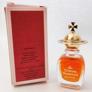May include: A Vivienne Westwood Boudoir perfume bottle with amber liquid and a gold orb-shaped cap featuring a cross, standing next to its rectangular pink and red cardboard box printed with ingredient details and manufacturer information.