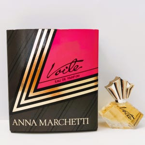 May include: A perfume bottle and box set. The box is black with gold and pink accents, and the words "Voile Eau de Parfum" are visible. The perfume bottle is a small, clear, geometric shape with a silver cap.