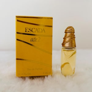 Puede incluir: Una caja de perfume rectangular amarilla con el texto ESCADA acte 2 y 4 ML e impreso en el frente, acompañada de un pequeño frasco de perfume de vidrio con una tapa cónica dorada y acanalada y un cuerpo transparente con líquido amarillo claro.