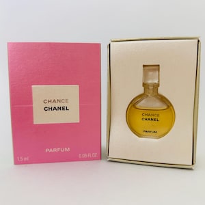 May include: A miniature perfume bottle and box set. The perfume bottle is round with a clear stopper, filled with a golden liquid. The box is pink with the words "Chance Chanel" and "Parfum" printed on it. The set is 0.05 fl oz.