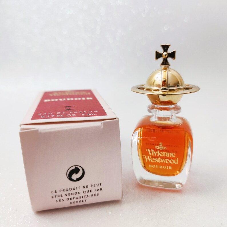 May include: A miniature Vivienne Westwood Boudoir eau de parfum bottle with an amber liquid and a gold orb-shaped stopper featuring a cross. Beside it sits a light pink box labeled 0.17 fl oz and 5 ml with red text and a recycling symbol.