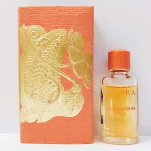 Op de afbeelding: Een vintage parfumflesje met een perzikkleurige doos met een gouden bloemenontwerp. De fles heeft een doorzichtige glazen body met een perzikkleurige dop en bevat een licht amberkleurige vloeistof. Het etiket luidt "SEORA" en "HAUTE COUTURIER PARIS."