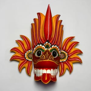 Pode incluir: Uma máscara de madeira pintada à mão com um rosto central, olhos pretos grandes, uma boca larga com dentes brancos e uma língua saliente. A máscara é decorada com padrões vibrantes de chamas vermelhas e amarelas e uma crista alta.