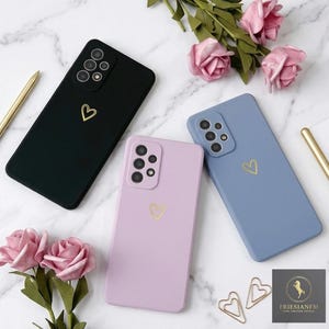 May include: Three smartphone cases in black, light purple, and dusty blue, each featuring a small gold heart outline on the back. The cases have a multi-lens camera cutout and are arranged alongside pink rose blossoms and gold-toned pens.