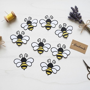 May include: A collection of handmade bee-shaped cutouts with black, yellow, and white details. The bees are arranged around a bundle of lavender, spools of thread, a tag that says "Handmade", and small scissors.