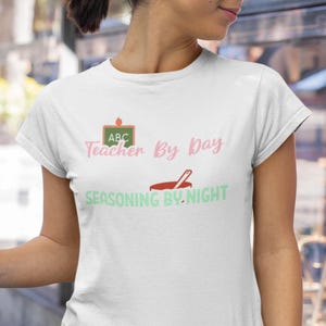 May include: White t-shirt with the text "Teacher By Day Seasoning By Night" in pink and green. The design includes an apple on a chalkboard and a cooking pot with a spoon. The shirt is a casual wear item.