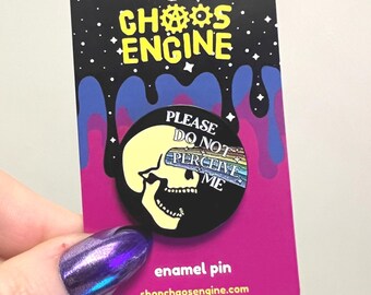 May include: Enamel pin with a skull design. The pin has a black background with a yellow skull and the text "PLEASE DO NOT PERCEIVE ME". The pin is on a pink card with the words "CHAOS ENGINE" and "enamel pin".