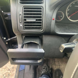 May include: A black, 3D-printed car cup holder installed in a vehicle's dashboard. The cup holder is rectangular with a curved opening and is positioned below the air vent. The car's interior is visible, including the dashboard and a portion of the steering wheel.