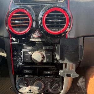 May include: A black car dashboard center console featuring two circular air vents with red trim, a central infotainment display, a radio control panel with buttons and a dial, and a black plastic phone mount attached to the right side.