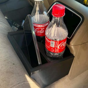 Pode incluir: Um organizador de consola de carro preto e retangular com dois compartimentos. Um compartimento contém uma garrafa de plástico transparente com tampa branca e rótulo vermelho Coca-Cola, o outro um smartphone e uma garrafa com tampa vermelha.