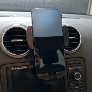 以下が含まれることがあります： 黒いスマートフォンが、ダッシュボードに取り付けられた車の電話ホルダーに固定されています。 ホルダーは黒と茶色で、ダッシュボードはグレーです。 車の通気口が電話の両側に表示されています。