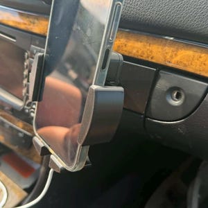 Puede incluir: Un soporte de plástico negro para smartphone fijado al salpicadero de un vehículo, que sostiene un teléfono en posición vertical. El soporte cuenta con brazos laterales ajustables y un soporte inferior, con un cable de carga blanco conectado.