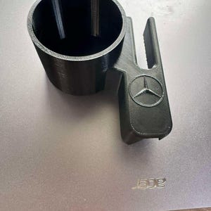 Puede incluir: Un portavasos negro para coche, impreso en 3D, con el logotipo de Mercedes-Benz. El portavasos tiene una abertura circular y un asa con el emblema de Mercedes-Benz. El artículo está diseñado para encajar en el interior de un coche.