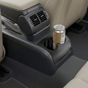 以下が含まれることがあります： カップホルダーと通気孔が内蔵された車のセンターコンソール。茶色の飲料缶がカップホルダーに置かれています。コンソールは黒色で木目調のトリム、座席はベージュ色です。