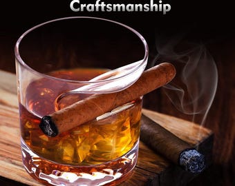May include: A rocks glass filled with amber liquid and ice, with a lit cigar resting on the rim. A second cigar is visible, along with the text "Traditional Handmade Craftsmanship". The scene is set on a wooden surface.