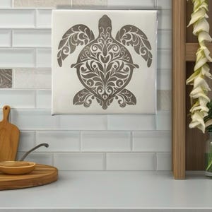 May include: A square, off-white tile featuring a detailed, gray turtle design. The turtle's shell and body are filled with intricate floral patterns. The tile is mounted on a white tiled wall, suggesting a decorative or functional kitchen element.