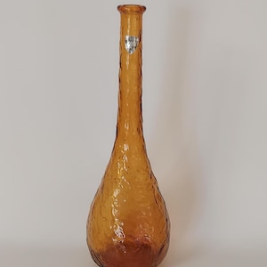May include: An amber-coloured glass vase with a textured surface. The vase has a long, slender neck and a bulbous base. A small, silver label is affixed near the top. The vase is likely vintage and suitable for displaying flowers or as a decorative piece.