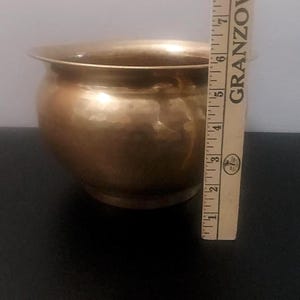 Pode incluir: Uma tigela redonda de latão martelado com uma borda larga e alargada. Uma régua de madeira está posicionada verticalmente ao lado da tigela, mostrando uma altura de aproximadamente 16,5 cm da base até a borda superior da tigela.