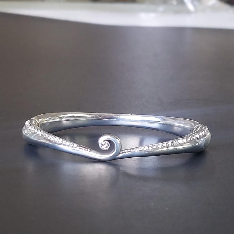 May include: A silver-toned hinged bangle bracelet featuring a polished finish and a central spiral design element. The band is accented with rows of small, clear embedded stones that follow the curved lines of the metal, creating a textured look.