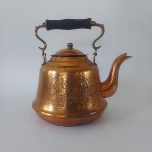 Puede incluir: Una tetera de cobre pulido con cuerpo redondeado, un patrón floral grabado en el frente, un pico curvo y un asa de metal articulada rematada con una empuñadura de madera marrón oscura y una tapa con un pomo central.