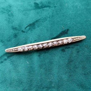 Può includere: Una lunga e sottile spilla a barra color oro con una fila di dieci strass trasparenti a taglio rotondo incastonati al centro. La montatura in metallo presenta una delicata texture perlata lungo i bordi superiore e inferiore.