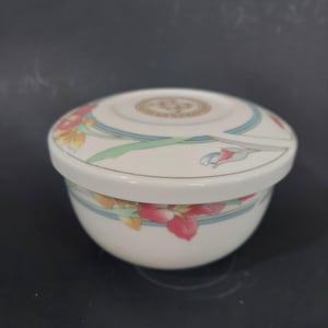 May include: A white ceramic trinket box with a lid. The box and lid are decorated with colorful floral patterns in pink, yellow, and blue. A gold emblem is centered on the lid.