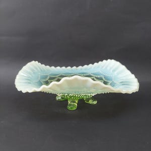 Può includere: Un piatto vintage in vetro opalescente con bordo a balze. Il piatto presenta un interno azzurro chiaro con un motivo delicato e poggia su tre piedini verdi strutturati. Il bordo è bianco.