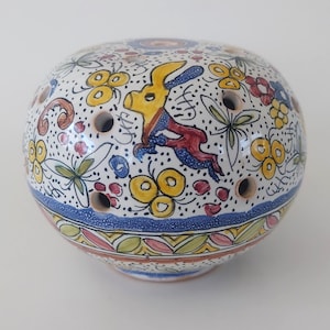 Peut inclure: Objet décoratif rond en céramique, à base blanche, orné de motifs floraux et de lapins colorés. Le design comprend des accents jaunes, rouges, bleus et verts. Il comporte plusieurs petits trous et une bordure bleue, mettant en valeur un style peint à la main.