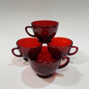 Könnte beinhalten: Vier rubinrote Teetassen aus Glas mit Henkeln sind pyramidenförmig gestapelt. Die Tassen haben eine abgerundete Schalenform und eine glatte, glänzende Oberfläche. Die Tassen sind auf einer weißen Oberfläche angeordnet.