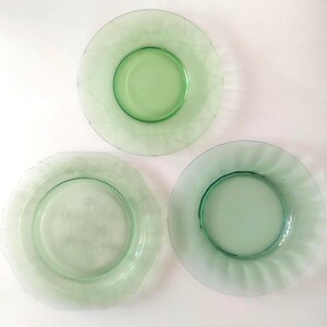 Op de afbeelding: Drie groene glazen borden in verschillende ontwerpen. Eén bord is rond met een gladde rand, een ander heeft een geschulpte rand en de derde heeft een golvende rand. De borden zijn lichtgroen.