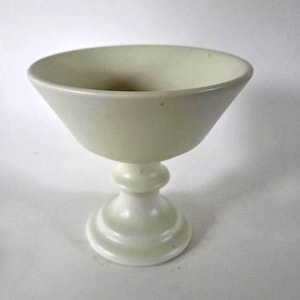 May include: A pale off-white pedestal bowl with a wide, flared rim and a smooth, matte finish. The vessel features a rounded, tapered body supported by a sturdy, circular base with a tiered, stepped design, creating a classic, balanced silhouette.