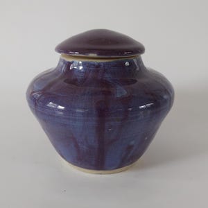 May include: A ceramic urn with a deep purple glaze and a matching lid. The urn has a rounded body and a slightly flared base. The lid is a flat, rounded shape. The glaze has a glossy finish with streaks of blue.