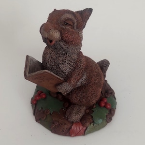 May include: A brown squirrel figurine reading a book with the text "Carols and Christmas". The squirrel is sitting on a green base with red berries and autumn leaves. The figurine has a detailed, textured appearance.