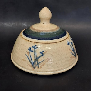 Puede incluir: Un tarro de cerámica beige con tapa y acabado moteado. La tapa presenta un pomo y una banda de rayas azules, verdes y marrones. Diseños florales azules están pintados en el exterior del tarro. El tarro es hecho a mano.