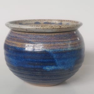 May include: A round ceramic bowl with a wide rim, featuring a speckled light grey top edge and a deep blue glazed body with horizontal ridges. The glaze transitions from a dark navy blue at the base to a lighter blue and tan hue near the top rim.