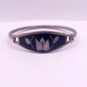 Peut inclure: Un bracelet jonc articulé de couleur argentée avec une incrustation ovale centrale en pierre noire. L'incrustation est ornée d'un motif floral composé de pièces de coquillage blanches et irisées disposées en pétales sur la surface sombre.
