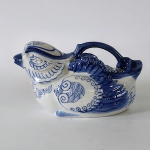 May include: A ceramic bird-shaped creamer or small pitcher. The white porcelain is decorated with blue floral and feather designs. It has a handle and a spout, and the bird's head is tilted slightly upward.