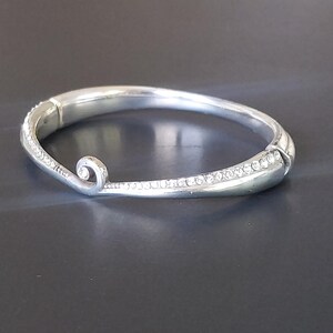 May include: A silver-toned hinged bangle bracelet featuring a smooth, polished finish and a curved, swirling end detail. The bracelet is accented with a row of small, clear, sparkling crystals embedded along the outer edge of the band.