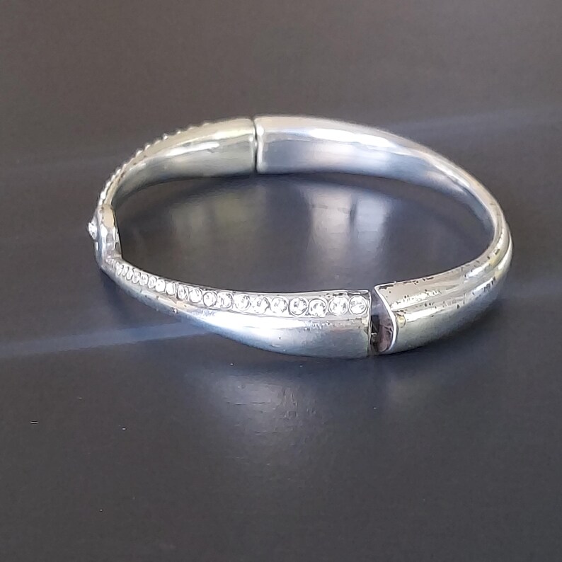May include: A silver-toned hinged bangle bracelet featuring a smooth, polished metal finish on one side and a curved section inlaid with a row of small, clear, sparkling crystals. The bracelet has a secure clasp mechanism and a rounded, solid design.