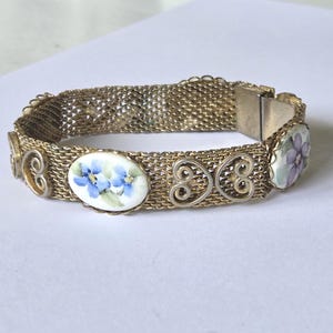 May include: A gold-tone mesh bracelet featuring alternating oval ceramic plaques painted with blue and purple floral designs, interspersed with decorative metal scrollwork elements and a secure metal clasp closure mechanism.