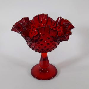 May include: A vibrant red glass compote dish featuring a raised hobnail texture across the bowl. The rim is wide and deeply ruffled with a scalloped edge. The bowl sits atop a smooth, tapered pedestal stem that connects to a circular base.