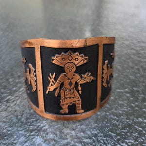 May include: A wide cuff bracelet featuring a metallic copper frame and a black textured inlay. The center of the bracelet displays a detailed copper-colored figure with a large headdress, flanked by additional geometric symbols on the sides.