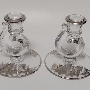May include: Two clear glass candle holders with silver-colored rims and floral designs on the base. The candle holders have a unique, curved design. The bases are round and have a silver floral pattern around the edges.