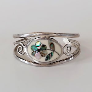 May include: Silver cuff bracelet with an oval centrepiece. The centrepiece features a flower design with iridescent abalone shell petals and a cream background. The bracelet has a double-band design with decorative swirls.