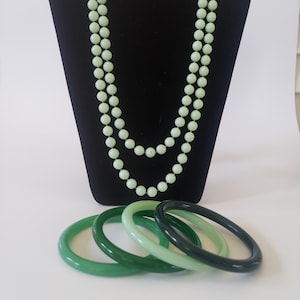 May include: A long, double-strand necklace featuring pale mint green spherical beads, accompanied by a set of four smooth, circular bangle bracelets in varying shades of green, ranging from light mint to a deep, dark forest green hue.