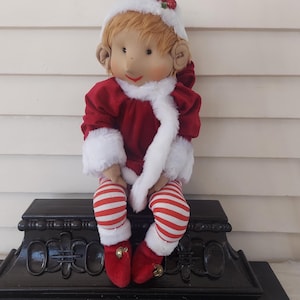Puede incluir: Muñeca de elfo navideño con atuendo rojo y blanco. La muñeca tiene una chaqueta de terciopelo rojo con ribete de piel blanca, medias a rayas rojas y blancas y zapatos de terciopelo rojo con cascabeles dorados. Lleva un gorro blanco con bayas rojas.