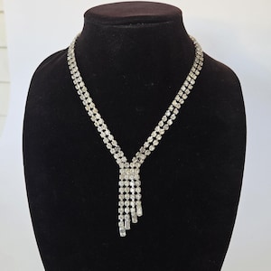 May include: A silver-toned necklace featuring two parallel rows of clear, faceted rhinestones that converge at a central rectangular link. From this link, four vertical strands of varying lengths hang down, each set with clear, faceted rhinestones.