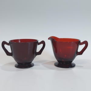 May include: A ruby red glass sugar bowl with two handles and a matching creamer with a spout. Both pieces have a rounded base and are set against a white background. The set is likely vintage.