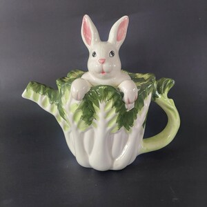 May include: A charming ceramic teapot shaped like a cabbage, with a white rabbit peeking out. The rabbit has pink ears and a friendly face. The teapot is white with green detailing, ideal for a spring or Easter-themed kitchen.