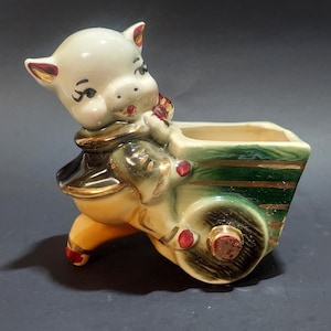 Puede incluir: Una maceta de cerámica vintage con forma de cerdo empujando un carro verde y dorado. El cerdo es de color crema con detalles en rojo y un contorno facial negro. El carro tiene una abertura rectangular.